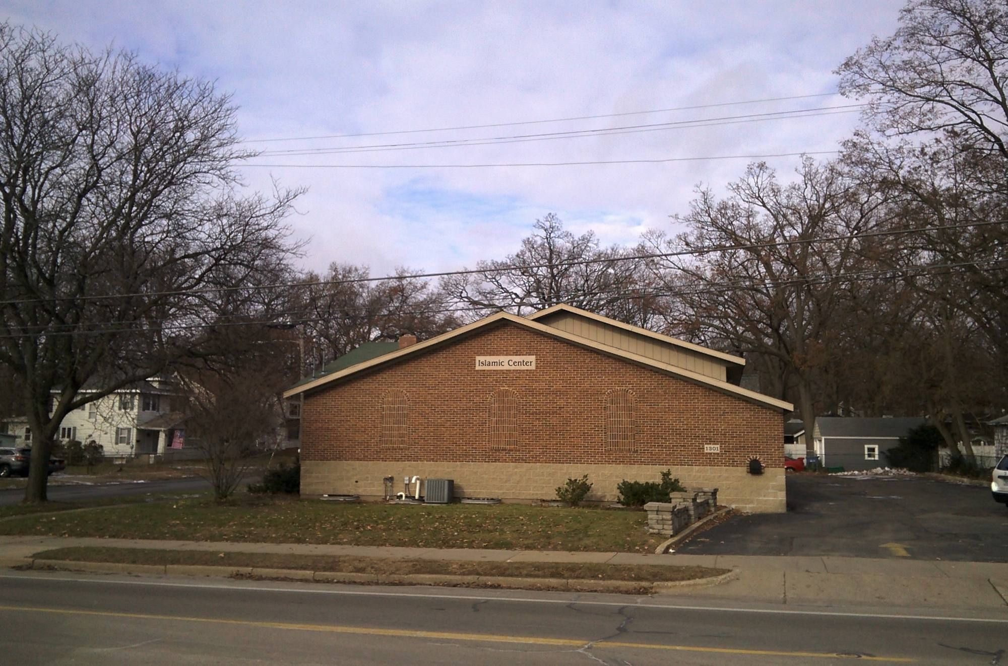A low single-storey brick building with a pitched roof behind a lawn and bare trees, beside a suburban road.