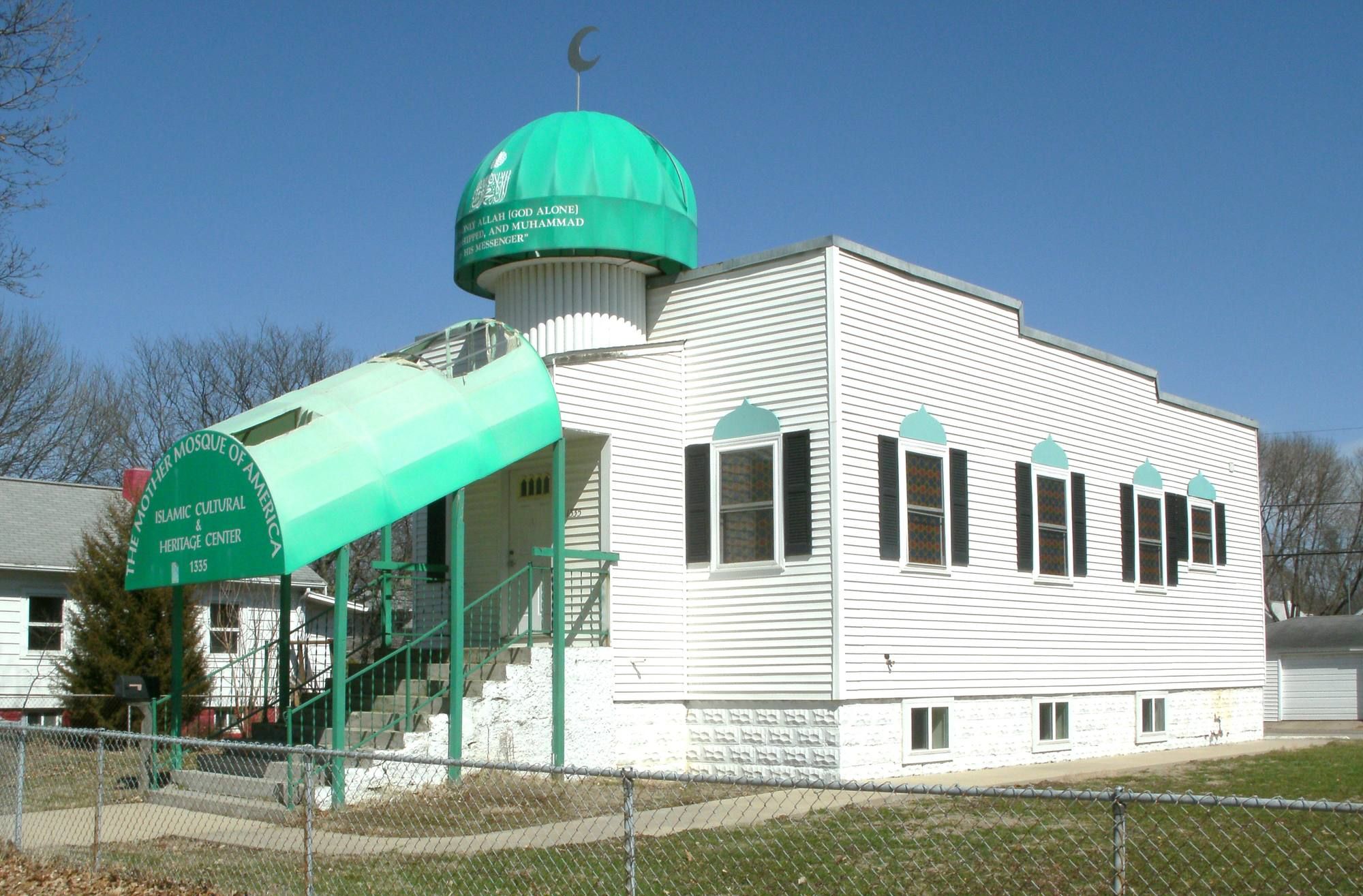 A small white clapboard building with a green dome and a green awning over the entrance steps, on a residential street under a blue sky.