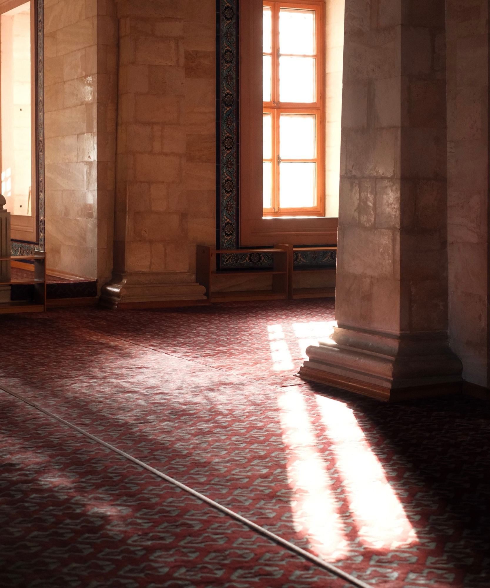 A tall window in a stone wall glowing with light, throwing long bright bars across a red patterned carpet beside a pillar.